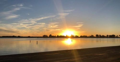 A serene sunset over a calm lake, with silhouettes of trees and poles reflected in the water.