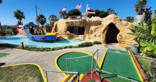 Colorful mini-golf course featuring a pirate ship, waterfall, and cave, surrounded by palm trees and blue sky.