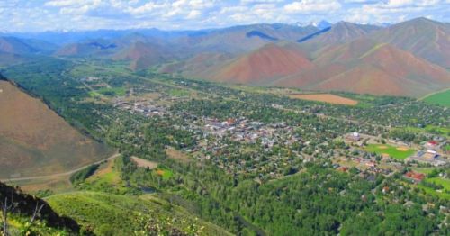 Aerial view of a small town surrounded by mountains and greenery, with a clear blue sky above.