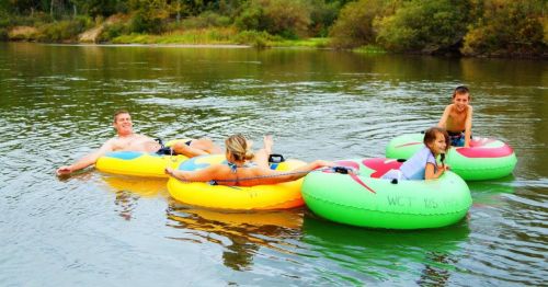 Four people relax on colorful inner tubes in a calm river, surrounded by greenery and trees.