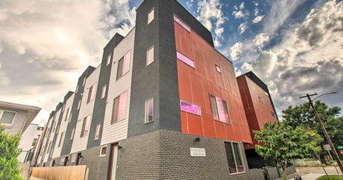Modern multi-story building with a mix of dark and light panels, large windows, and a cloudy sky in the background.