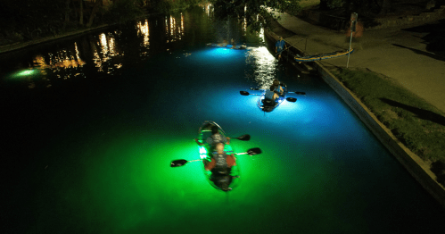 Two kayakers with illuminated paddles navigate a dark, reflective waterway at night.
