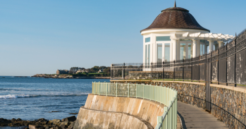 A scenic view of a coastal walkway with a gazebo and ocean waves under a clear blue sky.