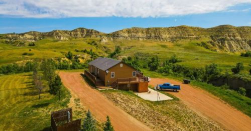 Aerial view of a brown house on a dirt road, surrounded by green hills and blue skies. A blue truck is parked nearby.