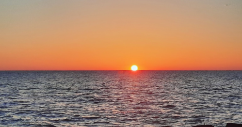 A vibrant sunset over calm ocean waters, with the sun dipping below the horizon and a gradient of warm colors in the sky.