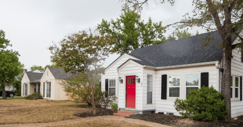 A white house with a red door, surrounded by greenery, alongside two other homes in a residential area.