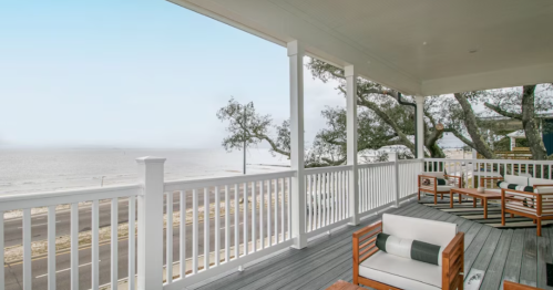 A coastal porch with wooden furniture overlooks a serene beach and calm sea under a clear sky.