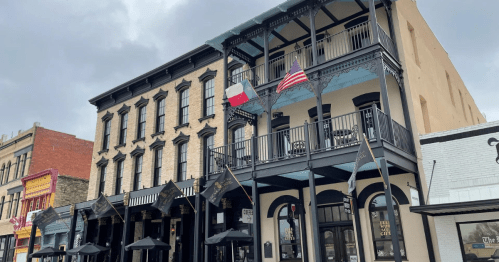 Historic building with balconies, featuring Texas and American flags, located in a vibrant downtown area.