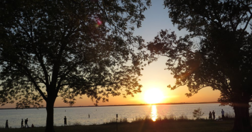 Sunset over a calm lake, framed by trees, with silhouettes of people enjoying the view along the shore.