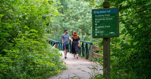 A couple walks on a trail surrounded by greenery, with a sign for Big Walnut Trail nearby.