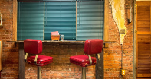 Two red bar stools at a rustic counter against a brick wall with blue blinds and exposed wood details.