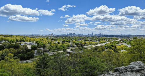 A panoramic view of a city skyline surrounded by lush greenery and rolling hills under a bright blue sky with clouds.