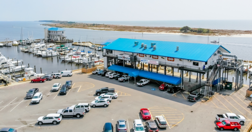Aerial view of a marina with boats, a blue-roofed building, and a parking lot filled with cars.