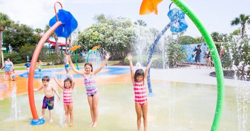 Children play joyfully in a splash pad, surrounded by colorful water features and greenery on a sunny day.