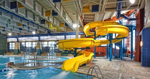Indoor water park featuring a large yellow waterslide, pool area, and seating along the edges. Bright and spacious atmosphere.