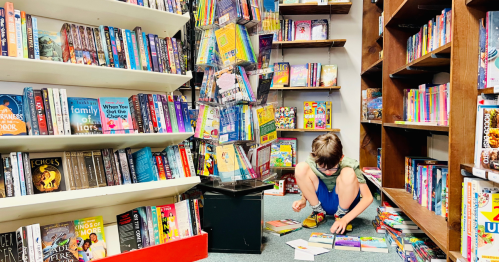 A child sits on the floor of a bookstore, surrounded by books, organizing a small stack of them.