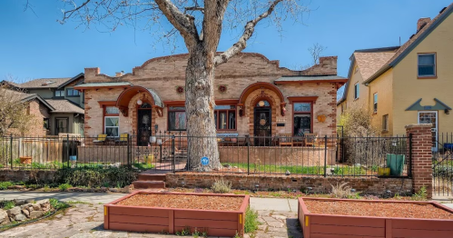 A historic brick house with arched doorways, surrounded by a garden and trees, under a clear blue sky.