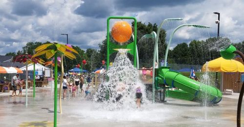 A vibrant water park scene with kids playing in splash pads, a water slide, and colorful umbrellas under a cloudy sky.