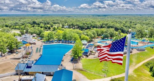Aerial view of a large swimming pool area surrounded by trees, with an American flag waving in the foreground.