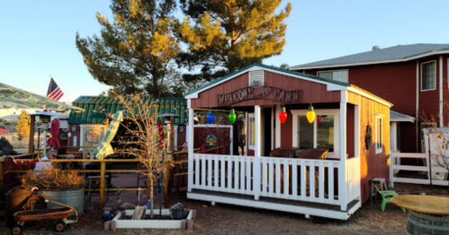 A colorful, rustic welcome hut with string lights, surrounded by trees and buildings in a sunny outdoor setting.