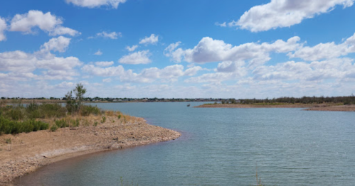 A serene river bends through a landscape of sandy banks and lush greenery under a bright blue sky with fluffy clouds.