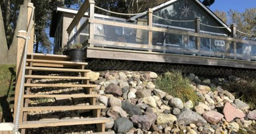 A wooden staircase leads up to a house with a glass railing, surrounded by rocks and greenery.