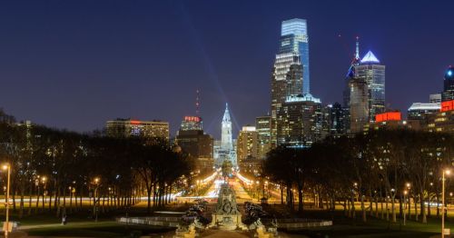 City skyline at night featuring illuminated buildings and a clear sky, with a street lined by trees in the foreground.
