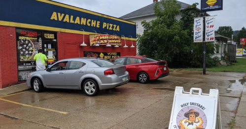 A gray car and a red car parked outside Avalanche Pizza, with a sign reading "Holy Shitake! It's Pizza Time!" nearby.