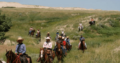 A group of cowboys and cowgirls riding horses through a grassy landscape with sand dunes in the background.