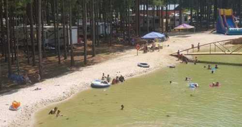 A sandy beach area by a lake, with people swimming, lounging, and enjoying activities under trees and tents.