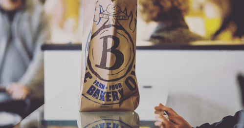 A brown paper bag with a large "B" logo, placed on a table, with a hand reaching towards it in a café setting.