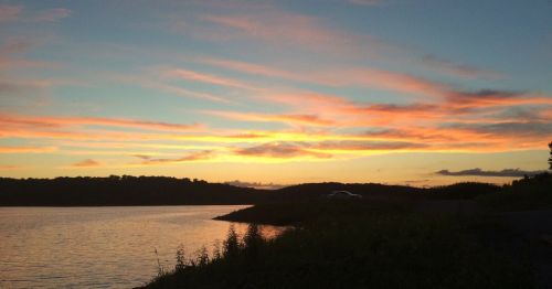 A serene sunset over a calm lake, with colorful clouds reflecting on the water and a silhouette of hills in the background.