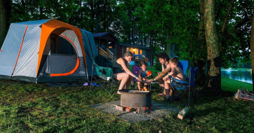 A family gathers around a campfire at night, with a tent and cabin in the background, enjoying time outdoors.