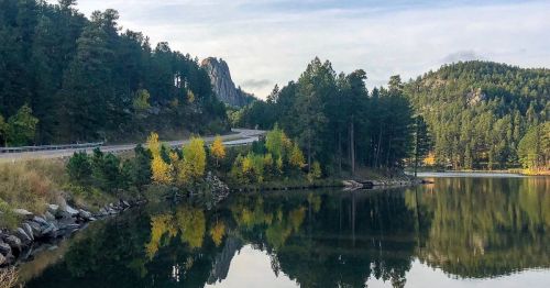 A serene landscape featuring a winding road, lush green trees, and a calm lake reflecting the surrounding mountains.