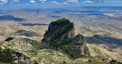 A rugged mountain peak rises above a vast, arid landscape under a blue sky with scattered clouds.