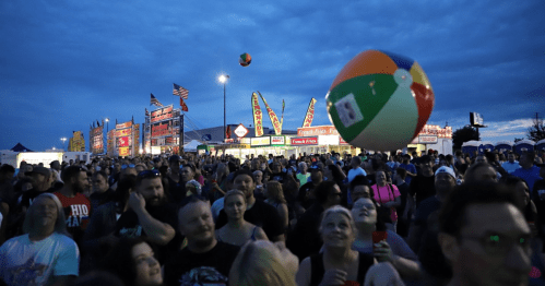 A large crowd at an outdoor event, with colorful food stalls and a beach ball flying overhead under a cloudy sky.