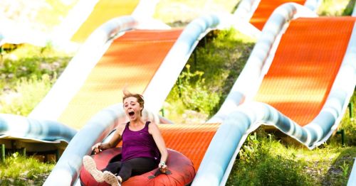 A woman joyfully slides down a colorful water slide on a tube, surrounded by green grass and vibrant slide paths.