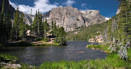 A serene mountain landscape featuring a clear lake, lush greenery, and towering rocky peaks under a blue sky.