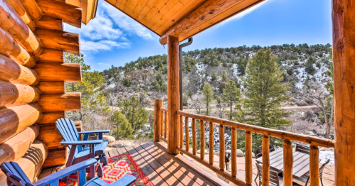 A cozy log cabin porch with two chairs, overlooking a scenic mountain landscape and trees under a clear blue sky.