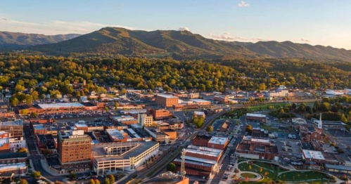 Aerial view of a town surrounded by mountains, featuring buildings, streets, and greenery under a clear sky.