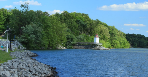 A serene lakeside scene featuring a lighthouse, rocky shore, and lush green trees under a blue sky.