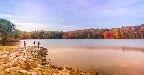 A serene lake surrounded by colorful autumn trees, with people walking along the rocky shore at sunset.