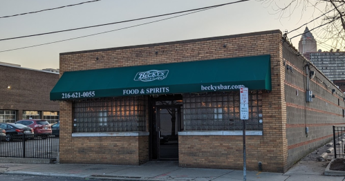 Exterior of Becky's Bar, featuring a green awning with the name and "Food & Spirits" signage. Brick building with parking nearby.