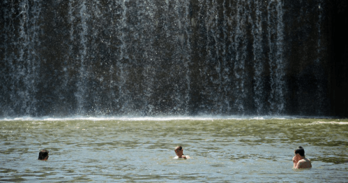 Three people wade in a pool beneath a large waterfall, surrounded by cascading water and a sunny atmosphere.