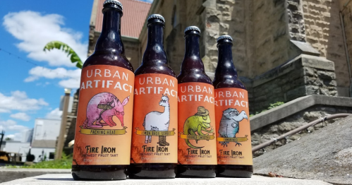 Four bottles of Urban Artifact beer, each with unique colorful labels, displayed outdoors against a stone wall.