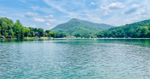 A serene lake surrounded by lush green trees and a mountain under a blue sky with fluffy clouds.
