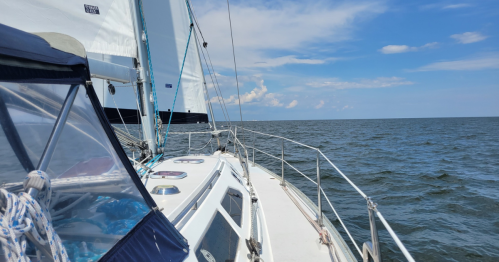 A sailboat glides over calm waters under a blue sky with scattered clouds.