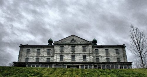 A large, old stone building with a gray sky overhead, set on a grassy hill.