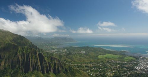 Aerial view of lush green mountains and coastline under a clear blue sky with scattered clouds.