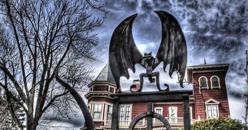 A large bat sculpture atop a gate, with a Victorian-style house and dramatic clouds in the background.